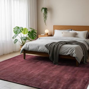 May include: A large, rectangular area rug in a deep, solid burgundy color with a soft, velvet-like texture. The rug is positioned on a light-colored floor, extending underneath a wooden bed frame, providing a bold contrast to the neutral bedroom decor.