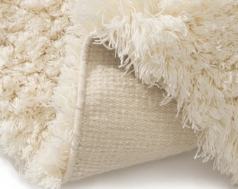 May include: A cream-colored shag rug with a high-pile, textured surface. The corner of the rug is folded over to reveal a woven, light beige fabric backing, showcasing the contrast between the soft, shaggy top and the structured underside.
