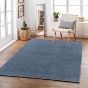Puede incluir: Una alfombra rectangular azul liso se extiende sobre un suelo de madera. La alfombra es de un azul apagado y polvoriento. La habitación incluye una maceta de cactus tejida, un espejo de cuerpo entero y una silla con un cojín gris. La alfombra mide aprox. 244 x 305 cm.