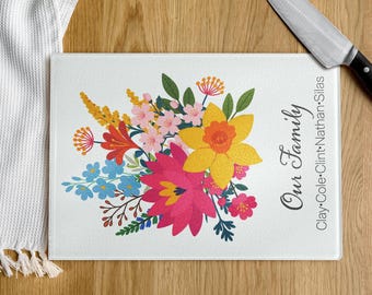 May include: A rectangular glass cutting board with a vibrant floral design. The bouquet features yellow, pink, red, and blue flowers. Text on the board reads "Our Family" and includes names. A knife rests on the wooden surface.