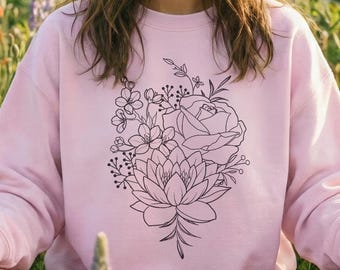 May include: A light pink sweatshirt featuring a black floral design. The sweatshirt has a crew neck and long sleeves. The floral design includes a rose, lotus flower, and other botanical elements. The person is sitting in a field of wildflowers.