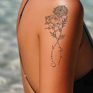 May include: Black floral tattoo design on the upper arm, featuring a peony and chrysanthemum. The tattoo includes the cursive text "Andressa" in a loop. The skin tone is light, and the background is blurred.