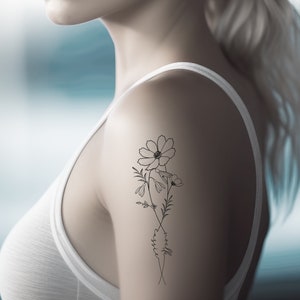 May include: A black and white floral tattoo design featuring cosmos flowers and delicate foliage. The tattoo includes the name "cassie" in cursive script. The design is on the upper arm of a person wearing a white tank top.