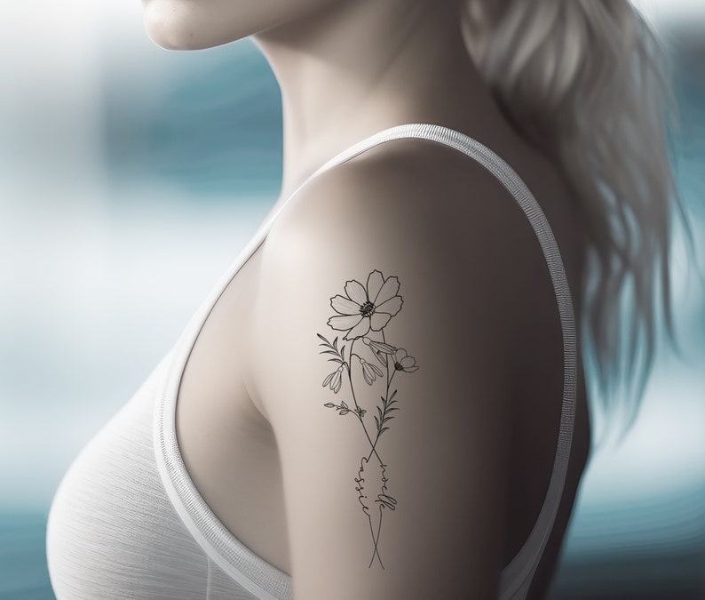 May include: A black and white floral tattoo design featuring cosmos flowers and delicate foliage. The tattoo includes the name "cassie" in cursive script. The design is on the upper arm of a person wearing a white tank top.