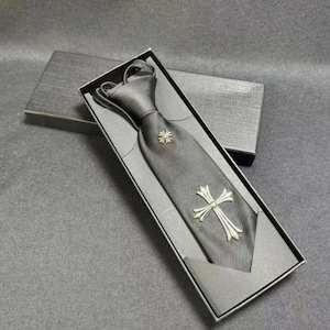 Puede incluir: Una corbata negra con textura de tejido diagonal, que presenta un pequeño emblema circular de tono plateado cerca del nudo y un adorno de cruz de tono plateado más grande colocado más abajo en la tela, presentada en una caja de regalo negra.