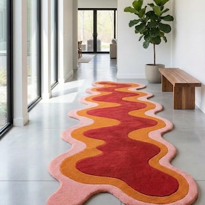 Puede incluir: Una colorida alfombra ondulada en un pasillo. La alfombra presenta un diseño en capas con tonos de rojo, naranja y rosa. Se coloca sobre un suelo gris, con un banco de madera y una planta en maceta en el fondo.