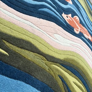 May include: A colorful rug with a water and koi fish design. The rug features shades of blue, green, pink, and orange. The design includes wavy lines to represent water and two orange koi fish swimming in the water.