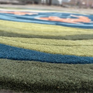May include: Close-up of a round rug with a textured surface. The rug features concentric bands of color, including shades of green, blue, and a hint of orange. The rug's design is reminiscent of a stylized landscape or abstract art.