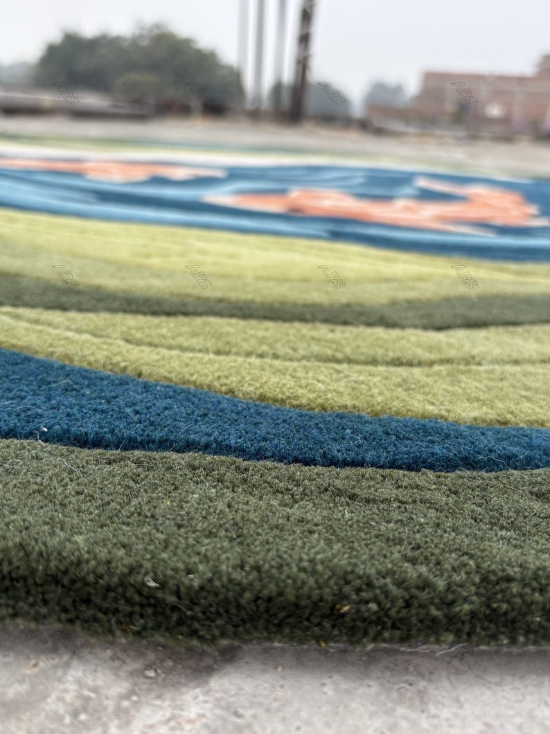 May include: Close-up of a round rug with a textured surface. The rug features concentric bands of color, including shades of green, blue, and a hint of orange. The rug's design is reminiscent of a stylized landscape or abstract art.