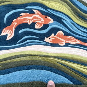 May include: Round rug featuring a koi fish design. The rug has a blue background with orange koi fish and green and blue wave patterns. The rug is made of a soft, textured material.