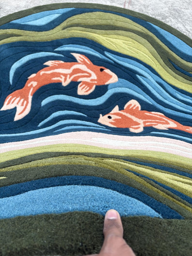 May include: Round rug featuring a koi fish design. The rug has a blue background with orange koi fish and green and blue wave patterns. The rug is made of a soft, textured material.