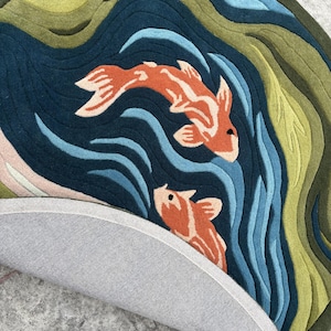 May include: Round rug featuring a koi fish design. The rug has a dark blue background with orange and white koi fish, blue water waves, and green and blue layered hills. The rug's underside is gray.