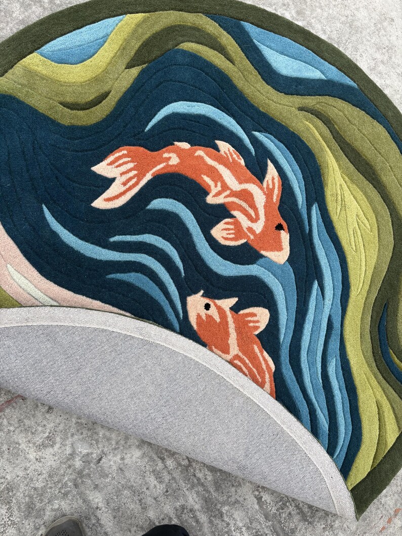 May include: Round rug featuring a koi fish design. The rug has a dark blue background with orange and white koi fish, blue water waves, and green and blue layered hills. The rug's underside is gray.