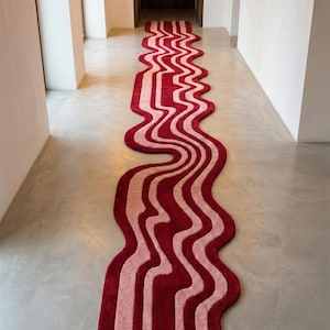 Könnte beinhalten: Ein langer, wellenförmiger Teppich mit einem einzigartigen Design. Der Teppich hat ein Muster aus abwechselnd roten und hellrosa Streifen, das eine fließende, abstrakte Ästhetik erzeugt. Der Teppich liegt in einem Flur mit weißen Wänden und Betonboden.