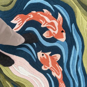 May include: A colorful rug featuring a koi pond design. Two orange and white koi fish swim in a blue pond with wave patterns. The rug has green, pink, and blue sections, creating a vibrant aquatic scene.