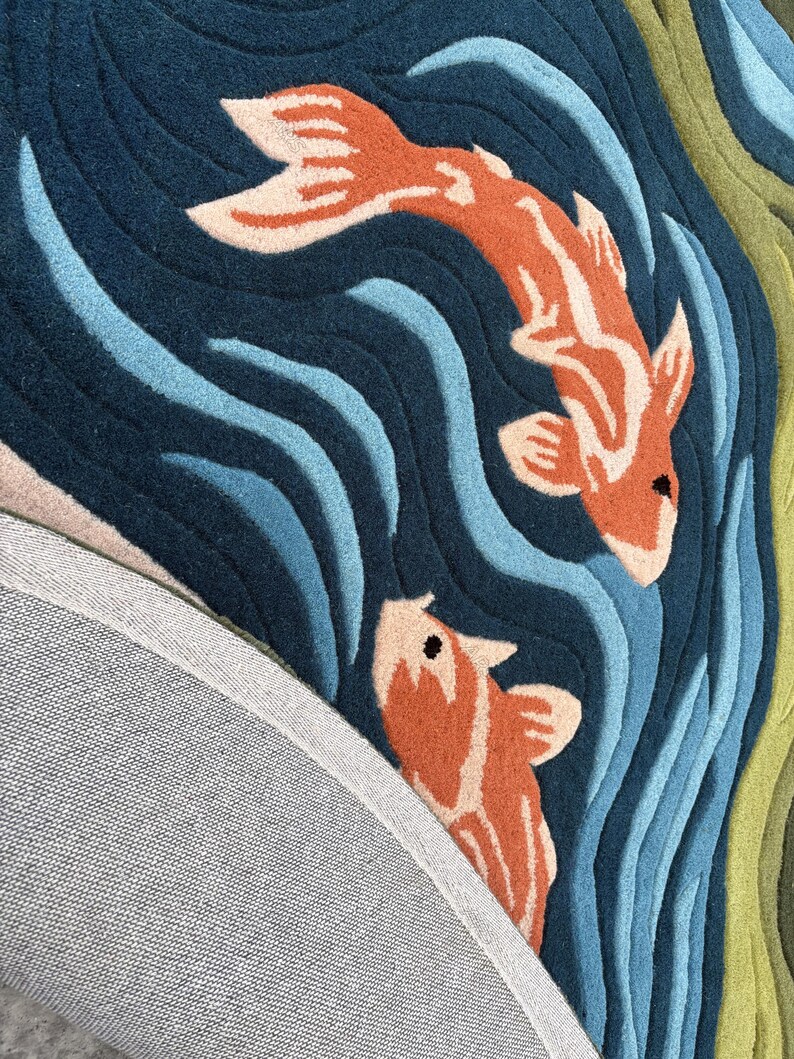 May include: A detailed rug featuring two orange and white koi fish swimming in a blue and teal water pattern. The rug has a dark blue background with wavy lines, and a gray backing is visible.