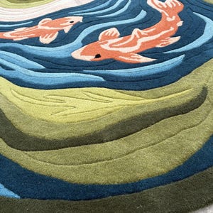 May include: A round rug featuring a koi fish design. The rug has a blue and green water pattern with two orange and white koi fish swimming across the surface. The rug is made of a soft, textured material.