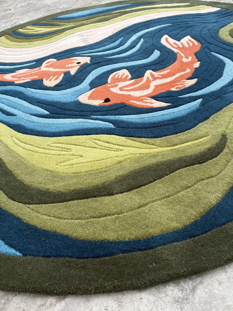 May include: A round rug featuring a koi fish design. The rug has a blue and green water pattern with two orange and white koi fish swimming across the surface. The rug is made of a soft, textured material.