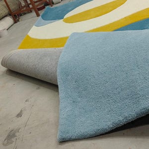 May include: A large area rug featuring a bold, abstract pattern with curved shapes in vibrant yellow, light blue, and dark teal. The rug has a thick, tufted texture and is partially folded, revealing a light grey, woven backing on the underside.