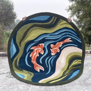 May include: Round rug featuring a koi pond design. The rug has a dark green border, with a blue and green water pattern. Two orange koi fish swim in the pond. The rug is held up by two hands.