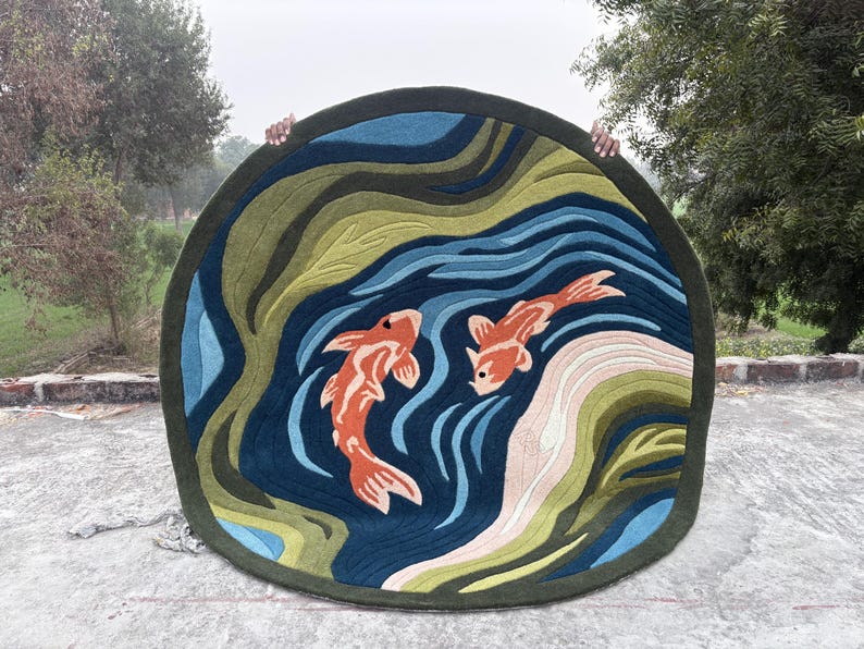 May include: Round rug featuring a koi pond design. The rug has a dark green border, with a blue and green water pattern. Two orange koi fish swim in the pond. The rug is held up by two hands.