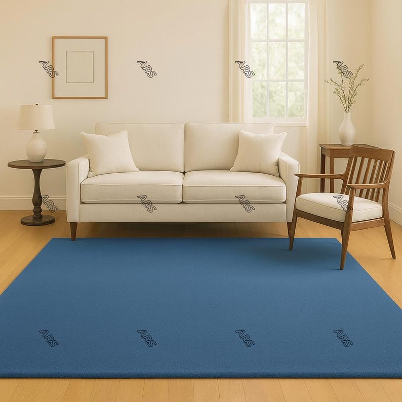 May include: A rectangular, solid blue rug covers the floor in a living room setting. A cream-colored sofa with matching pillows sits against a light-colored wall. A wooden chair and side table are also present.