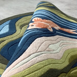 May include: A rolled-up, round rug with a koi fish design. The rug features a layered landscape with shades of blue, green, and beige. The koi fish is orange and white. The rug is made of a soft, textured material.
