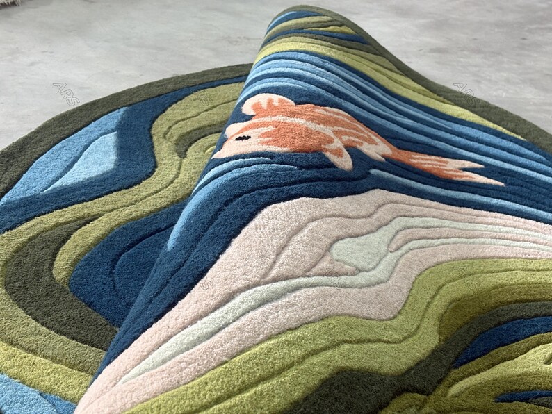 May include: A rolled-up, round rug with a koi fish design. The rug features a layered landscape with shades of blue, green, and beige. The koi fish is orange and white. The rug is made of a soft, textured material.
