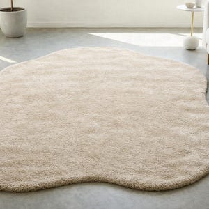 Op de afbeelding: Een beige, onregelmatig gevormd vloerkleed met een zachte, pluche textuur. Het kleed ligt op een betonnen vloer en de neutrale kleur past bij het minimalistische decor van de kamer. Afmetingen: ongeveer 213 x 152 cm.
