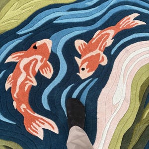 May include: A colorful rug featuring a koi pond design. The rug has a dark blue background with orange and white koi fish swimming in the water. The water is depicted with shades of blue, green, and pink.