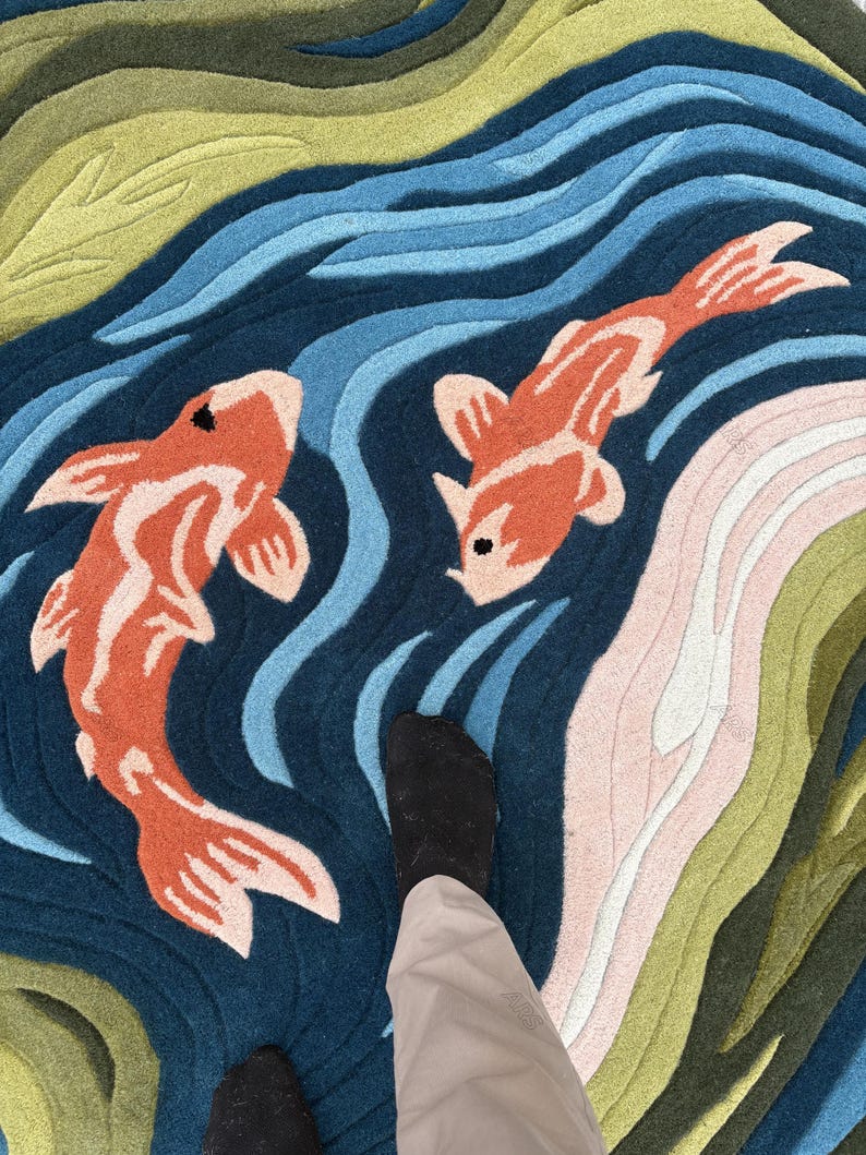 May include: A colorful rug featuring a koi pond design. The rug has a dark blue background with orange and white koi fish swimming in the water. The water is depicted with shades of blue, green, and pink.