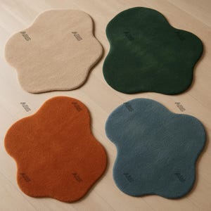 Op de afbeelding: Vier onregelmatig gevormde vloerkleden in beige, donkergroen, oranje en blauwgrijs. Elk kleed heeft een zacht, getextureerd oppervlak en een organische vorm. De kleden liggen op een lichte houten vloer.