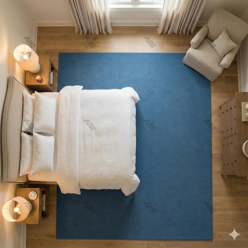 May include: An overhead view of a bedroom with a large blue rug, a bed with white bedding, and a beige armchair. Two bedside tables with lamps flank the bed. The room has hardwood floors and neutral-colored curtains.