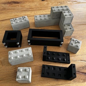 Könnte beinhalten: Eine Anordnung von Betonblöcken und schwarzen Metallrahmen auf einer Holzoberfläche. Die grauen Betonblöcke haben eine strukturierte Oberfläche. Die schwarzen Metallrahmen sind rechteckig und mit Schrauben versehen. Einige Blöcke haben Metallringe.