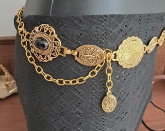 Pode incluir: Cinto de metal dourado com detalhes ornamentados. Apresenta um oval central com uma pedra preta, conectado por correntes a outros elementos decorativos, incluindo um pingente de moeda. O cinto é exibido em uma superfície preta.