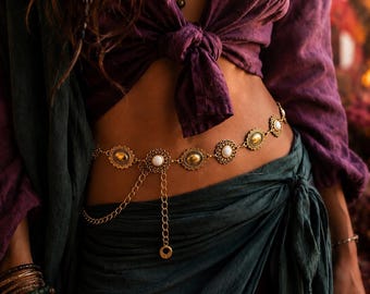 Pode incluir: Uma corrente de cintura dourada com uma série de elos circulares e ovais com bordas recortadas ornamentadas. Os elos são cravejados com cabochões dourados lisos e pedras brancas alternadas. Uma corrente fina e pendente pende do centro.