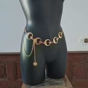 Peut inclure: Ceinture céleste dorée avec des croissants de lune, des étoiles et un pendentif soleil pendant. La ceinture est présentée sur un mannequin noir, mettant en valeur son design. La chaîne de la ceinture mesure environ 76 cm.