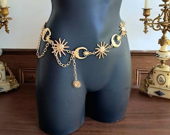 May include: Gold-colored belt featuring sun and crescent moon charms. The celestial-themed belt is worn on a black mannequin. The belt has a chain and a dangling sun charm. The belt is a decorative accessory.