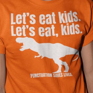 May include: An orange t-shirt with the text "Let's eat kids. Let's eat, kids." and a white dinosaur graphic. The phrase "PUNCTUATION SAVES LIVES" is printed below the graphic. The person is wearing yellow sunglasses.