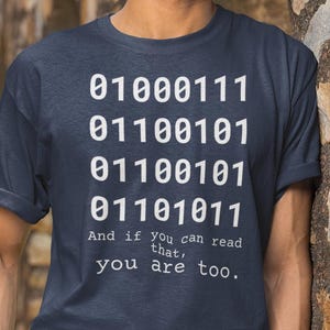 Op de afbeelding: Marineblauw t-shirt met binaire code en de tekst "And if you can read that, you are too." Het shirt wordt gedragen met een witte broek en een bruine riem. De persoon draagt een zwarte bril.