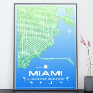 Op de afbeelding: Een ingelijste print van een Miami-kaart met groene en blauwe gradiënten. De kaart toont de indeling van de stad, met de tekst "MIAMI" en "WHERE THE OCEAN SHINES BRIGHT" onderaan, vergezeld van illustraties van palmbomen, flamingo's en een vogel.