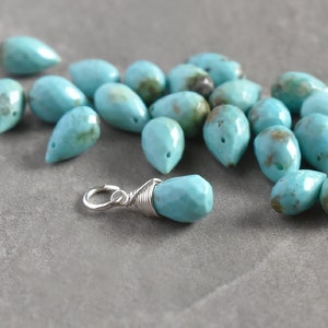 May include: A close-up of a turquoise gemstone charm, wrapped in silver wire, with a silver jump ring. The charm is surrounded by a scattering of turquoise stones in various shapes and sizes.