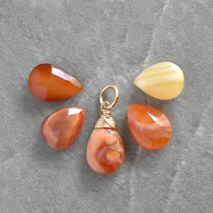 May include: A gold-colored pendant with a teardrop-shaped, orange-toned stone, surrounded by four loose, similarly colored stones. The stones are displayed on a gray, textured surface. The pendant is a charm.