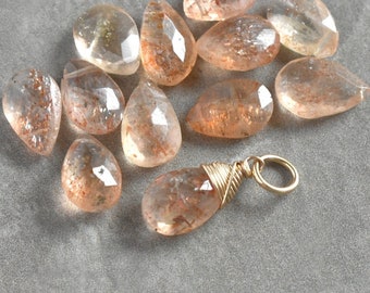 May include: A collection of 10 sunstone gemstone charms, each with a teardrop shape and a wire-wrapped loop for hanging. The stones are a light peach color with a shimmery, iridescent quality. One charm is shown with a gold-toned wire wrap and a jump ring.