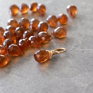 May include: A group of 20 brown gemstone beads in a teardrop shape, scattered on a light gray surface. One bead is attached to a gold wire loop.