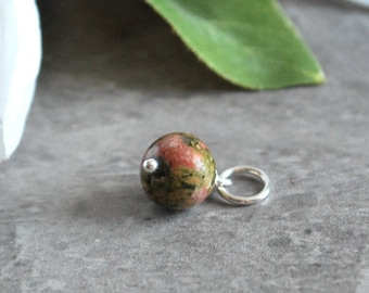 May include: A small, round unakite gemstone bead with a silver-coloured bail. The bead is a mix of green, pink, and brown colours. The bead is shown against a grey background with a white flower and green leaf.