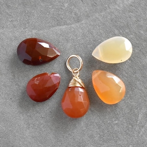 May include: A collection of gemstone charms and loose stones in shades of brown, orange, and cream. One charm features a gold-colored bail and wire wrapping. The stones are teardrop and oval shapes, displayed on a gray surface.