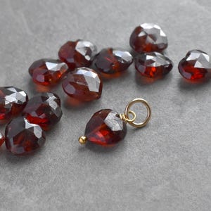 May include: A selection of faceted, heart-shaped, red-brown gemstones. One gemstone is attached to a small gold-coloured charm. The stones are scattered on a grey surface. The text at the bottom reads: (For 1 finished charm. Stone randomly selected. Enlarged to show detail).