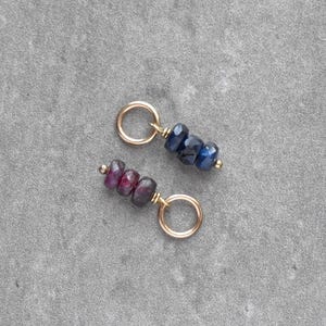 May include: Two gold-colored charms, each with a small loop and a cluster of gemstones. One charm features dark blue stones, the other has deep red stones. The charms are displayed on a gray surface.