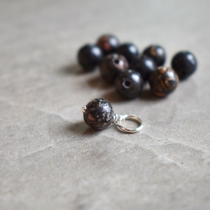 May include: Close-up view of a single dark brown and black speckled stone bead pendant on a silver chain, accompanied by a pile of similar beads in varying shades of dark brown and black with hints of reddish-brown. The beads appear to be made of natural stone or a similar material, exhibiting unique patterns and textures.  The pendant showcases the same speckled pattern as the other beads, suggesting a cohesive jewelry set or collection of handcrafted accessories.